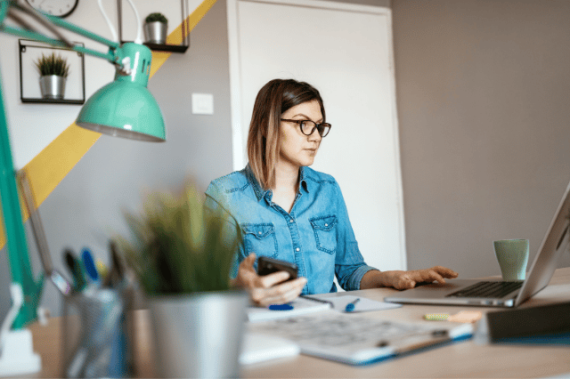 Woman working from home sitting at desk with laptop & phone in hand
