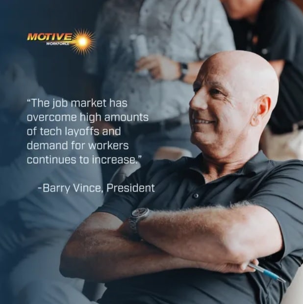 Barry Vince, President of Motive Workforce, sitting with arms crossed with people behind him